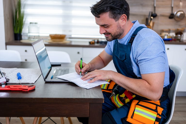 Handyman documents household repairs while seated at a kitchen table with a laptop and tools.