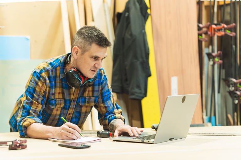 Male carpenter writing notes while working on laptop in woodworking shop