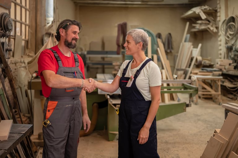 Two carpenters, a man and a woman, shaking hands and smiling in their busy wood workshop, symbolizing business partnership, agreement, and traditional craftsmanship